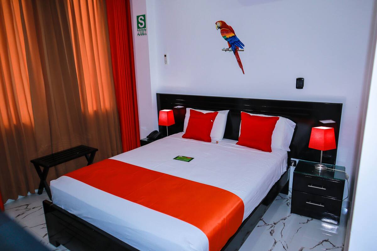 A neatly made bed with a red and white bedspread, a nightstand with a lamp, and a wall-mounted artwork of a parrot.