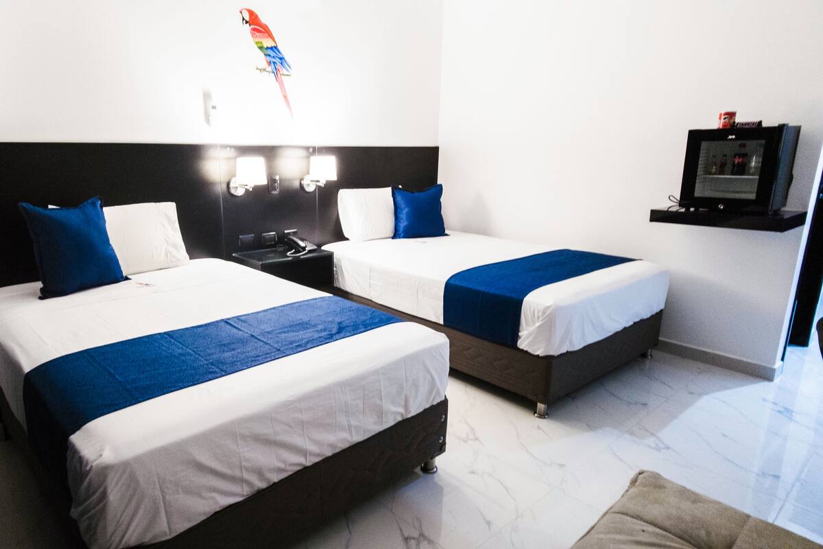 A hotel room with two beds, each with white bedding and blue accents, a black headboard, a small desk with a telephone, and a TV mounted on the wall.