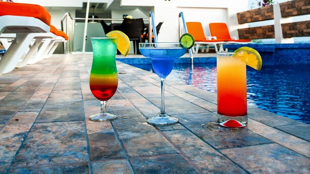 Un área junto a la piscina con tres bebidas coloridas, un edificio blanco con un letrero que dice "HOTEL PAPAGAYO" y sillas reclinables naranjas.