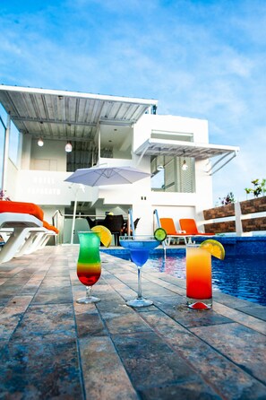 Un área junto a la piscina con tres bebidas coloridas, un edificio blanco con un letrero que dice "HOTEL PAPAGAYO" y sillas reclinables naranjas.