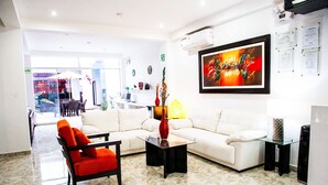 Un moderno vestíbulo de hotel con un sofá blanco, un sillón rojo y una mesa de centro de vidrio. Hay un cuadro abstracto grande en la pared y una unidad de aire acondicionado montada sobre el sofá.