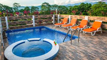 Une piscine avec un jacuzzi, entourée de chaises longues orange, le tout se fondant dans un décor de verdure luxuriante et de montagnes.