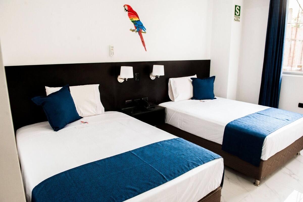 A hotel room with two beds, white bedding, blue accents, a black headboard, a wall-mounted lamp, and a colorful parrot decal.