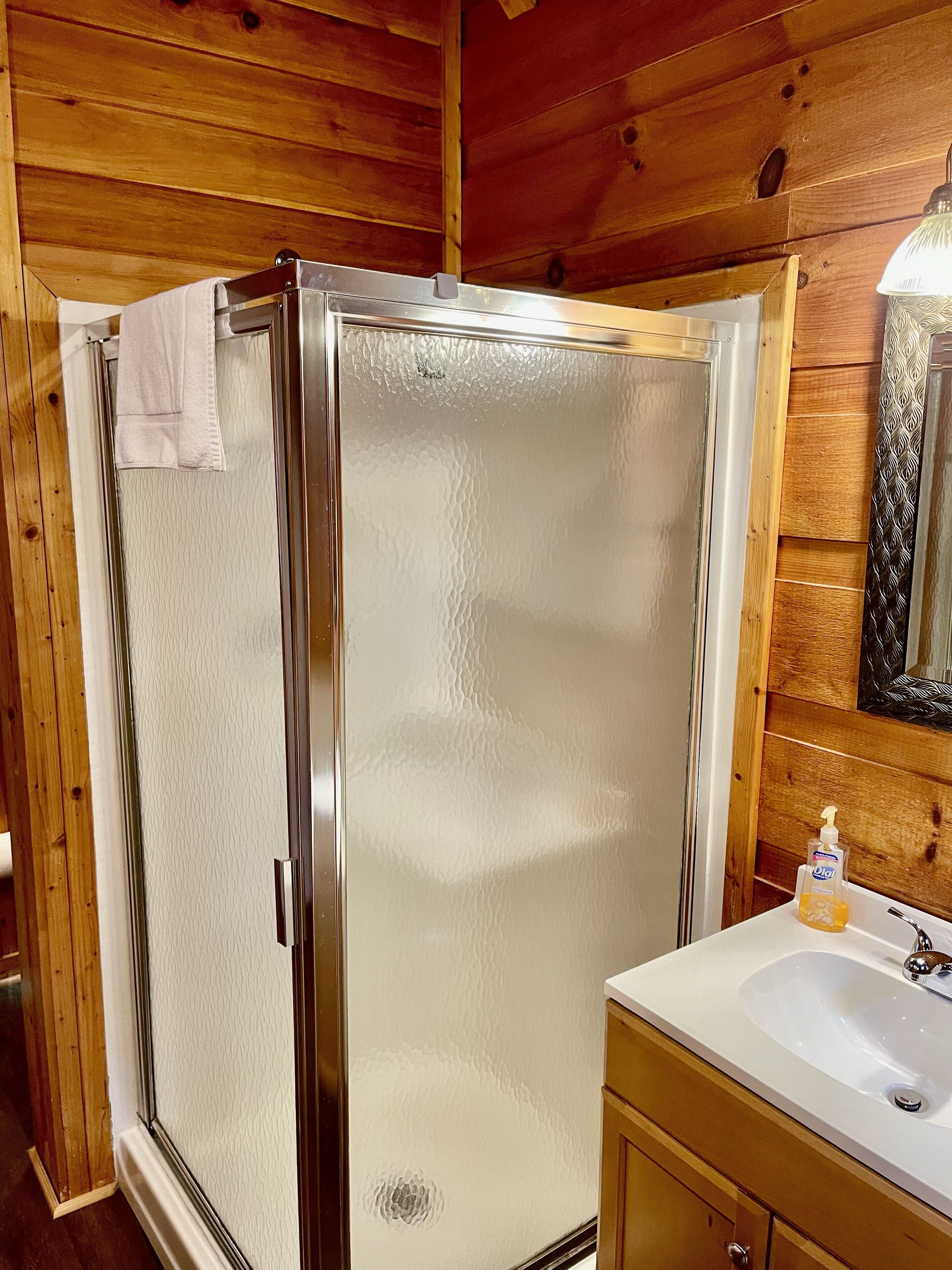 Une salle de bain rustique, avec une cabine en bois, une douche en verre, un lavabo blanc et un meuble en bois.