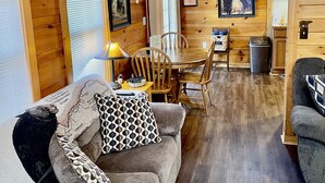 Ein gemütliches Blockhaus-Interieur mit Holzbalkendecke, einem Essbereich mit Tisch und Stühlen, einer Couch mit gemusterten Kissen und einem gerahmten Bild an der Wand.