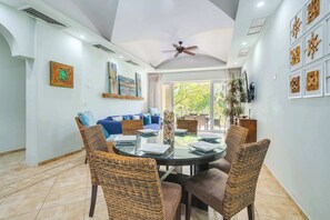 Un coin repas avec une table ronde en verre, des chaises en osier, un ventilateur de plafond et une vue sur l’extérieur.