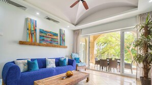 A living room with a blue sofa, a wooden coffee table, and a large window with a view of greenery. - Matapalo 201 | Steps from Tamarindo Beach (Tamarindo)