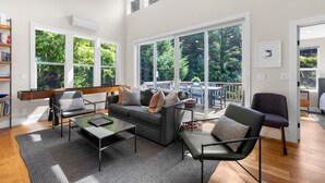 A modern living room with a sofa, armchairs, and a coffee table. There are large windows with a view of greenery outside.
