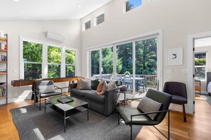 A modern living room with a sofa, armchairs, and a coffee table. There are large windows with a view of greenery outside.
