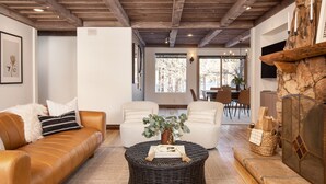 Una sala de estar con un sofá de cuero marrón, dos sillones blancos, una mesa de centro redonda de mimbre con un jarrón y objetos decorativos, y una chimenea de piedra con una protección de vidrio.