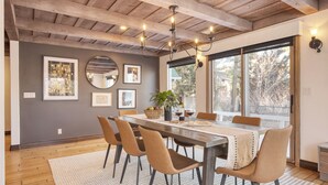 Un comedor con una mesa de madera, cuatro sillas, una araña, obras de arte enmarcadas y un ventanal con vistas.
