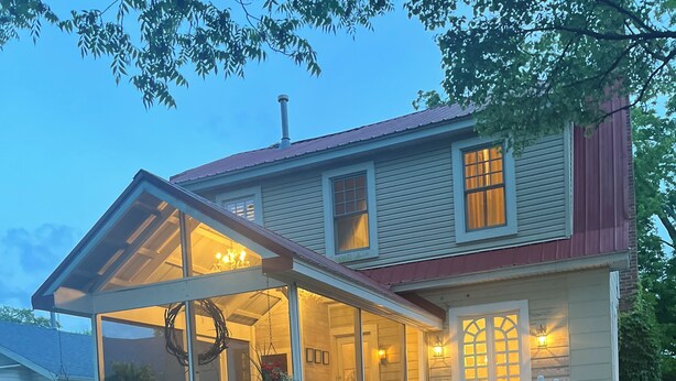 Een tweelaags huis met een overdekte veranda, een groot raam en een schoorsteen.
