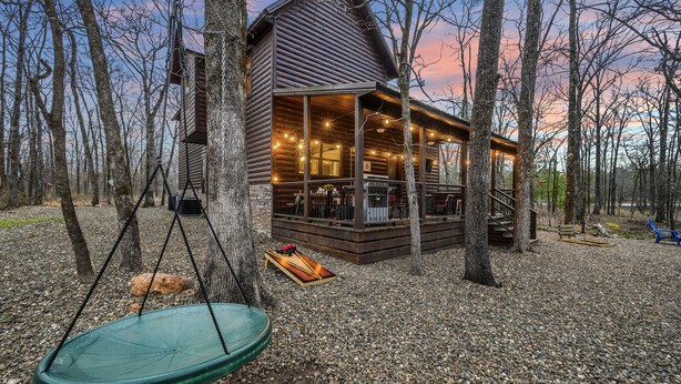 Une cabane avec une véranda, entourée d’arbres, et une chaise suspendue.