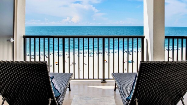 Un balcon avec deux fauteuils en osier, une vue sur une plage parsemée de parasols et un ciel dégagé.