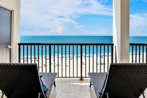 A balcony with two wicker chairs, a view of a beach with umbrellas, and a clear sky. - The Palms 1014 | Spacious 2 bedroom OBA beach front condo! (Orange Beach)