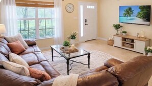 Un salon avec un canapé en cuir marron, une table basse en verre, une porte blanche, un téléviseur et une horloge murale.