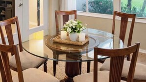 Une table de salle à manger ronde en verre, accompagnée de quatre chaises en bois, un set de table en fibres tressées avec des bougies et un vase rempli de fleurs.