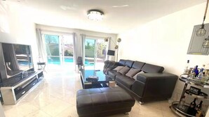 Un salon spacieux avec un grand canapé en cuir noir, une table basse en verre et un téléviseur à écran plat. Une porte-fenêtre coulissante donne accès à un balcon avec une piscine.