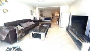 Un salon moderne avec un canapé, une table basse, un téléviseur et un coin cuisine à l’arrière-plan.