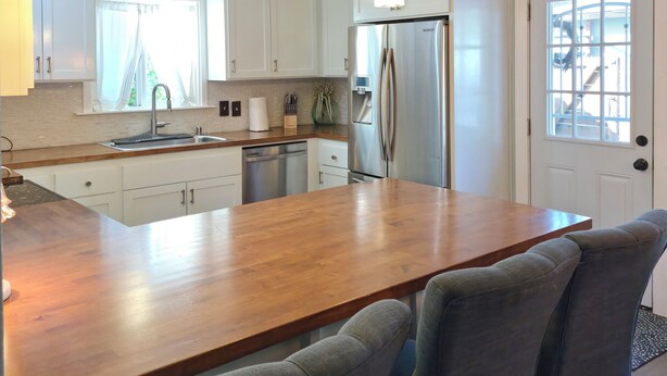 A modern kitchen with white cabinets, a stainless steel refrigerator, a wooden island with bar stools, pendant lights, and a window with curtains.