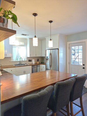A modern kitchen with white cabinets, a stainless steel refrigerator, a wooden island with bar stools, pendant lights, and a window with curtains. - Garden Clara (Yuba City)