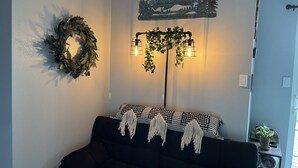 Olohuone, jossa on musta sohva, koristeellinen seppele, seinään kiinnitetty valaisin ja seinätaideteos.