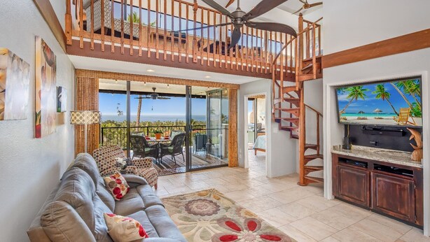 A spacious living area with a sofa, a TV, and a staircase leading to a balcony with a view of the ocean. - 🏖️Ocean View Condo + Pickleball | Top Floor w/ AC (Kailua Kona)