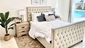 Une chambre à coucher avec une tête de lit capitonnée, une table de chevet avec une lampe, une grande plante en pot et une vue sur une piscine par la fenêtre.