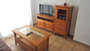 Een kleine woonkamer met een houten salontafel, een televisie op een houten kast en een tafel met een glazen blad.