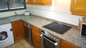 Een keuken met granieten aanrechtbladen, houten kastjes, een inbouwoven, een vaatwasser en een raam met zonwering.