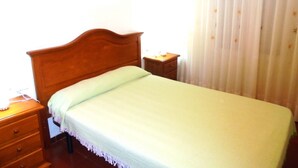 Een enkel bed met houten hoofdeinde in een kamer met nachtkastje, lade en klein tafeltje. Er is een raam met gordijnen.