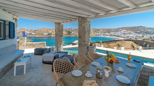 Zona pranzo all'aperto con vista sul mare e sulle colline, caratterizzata da pilastri in pietra, sedie in vimini e un tavolo apparecchiato per la colazione.