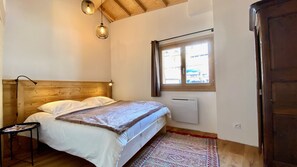 Um quarto com cabeceira de madeira, cama com roupa de cama branca, uma mesinha com revistas, uma janela com cortinas e um radiador.