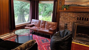 Una sala de estar acogedora con un sofá de cuero, una mesa redonda de centro y un sillón orejero negro. Se ve una chimenea de ladrillo y un cuadro de un paisaje montañoso.