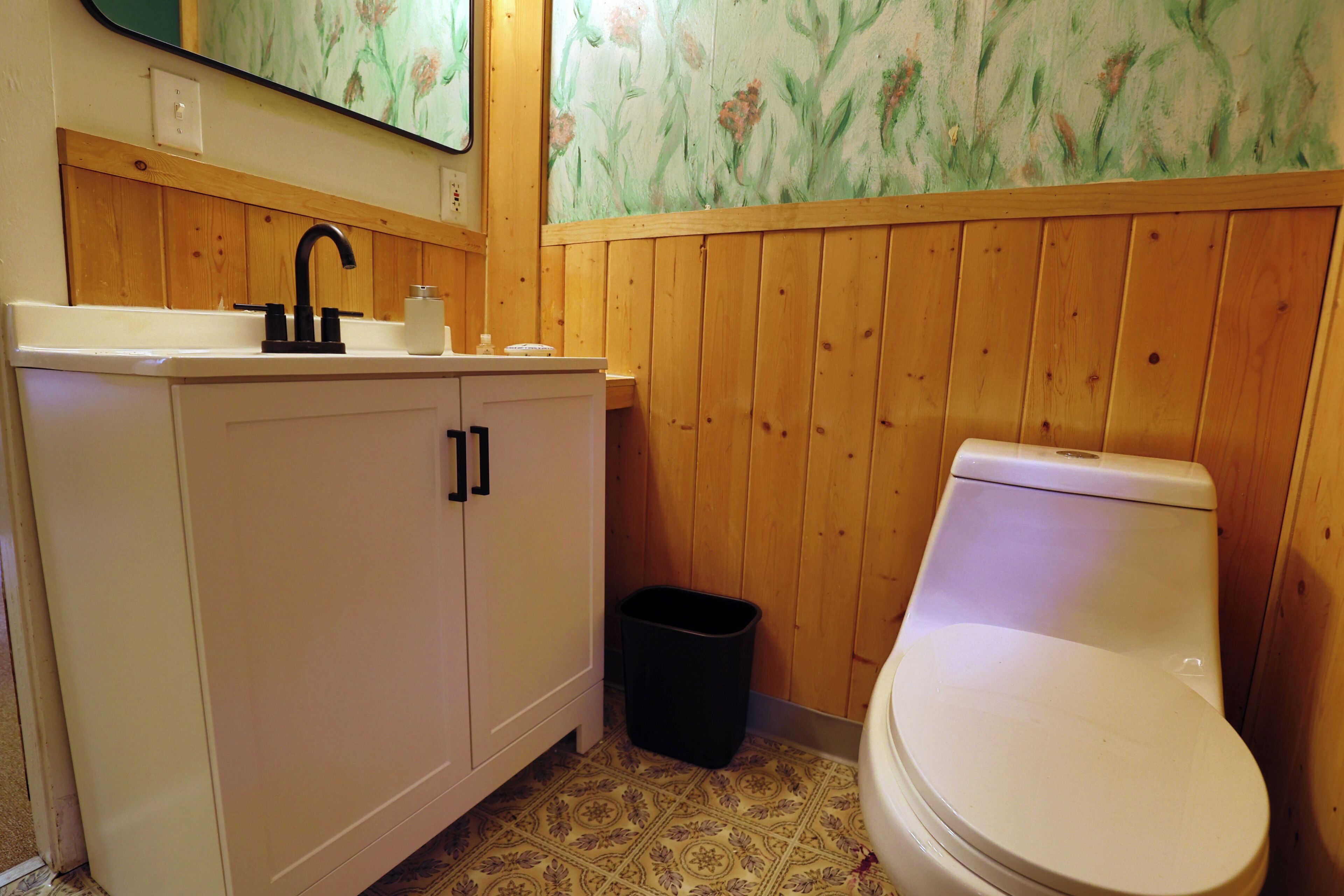 A bathroom with a wooden panel wall, a white sink with a black faucet, a white toilet, and a trash bin.