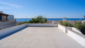 Eine Dachterrasse mit Blick auf das Meer, ein weißes Gebäude und etwas Grün.