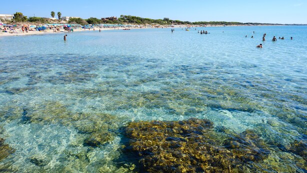Ein Strand mit klarem Wasser, badenden Personen und Sonnenschirmen im Sand.