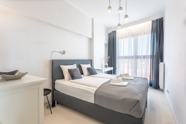 Une chambre d’hôtel moderne avec un grand lit, une table de chevet, un bureau et une vue sur la ville à travers des rideaux transparents.