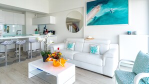 Un salon moderne avec un canapé blanc, une table basse ornée de fleurs, un coin cuisine et un grand tableau représentant une scène de plage.