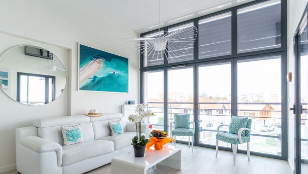 Un salon moderne avec un canapé blanc, une table basse ornée de fleurs, une grande fenêtre donnant sur un beau paysage et un ventilateur de plafond.
