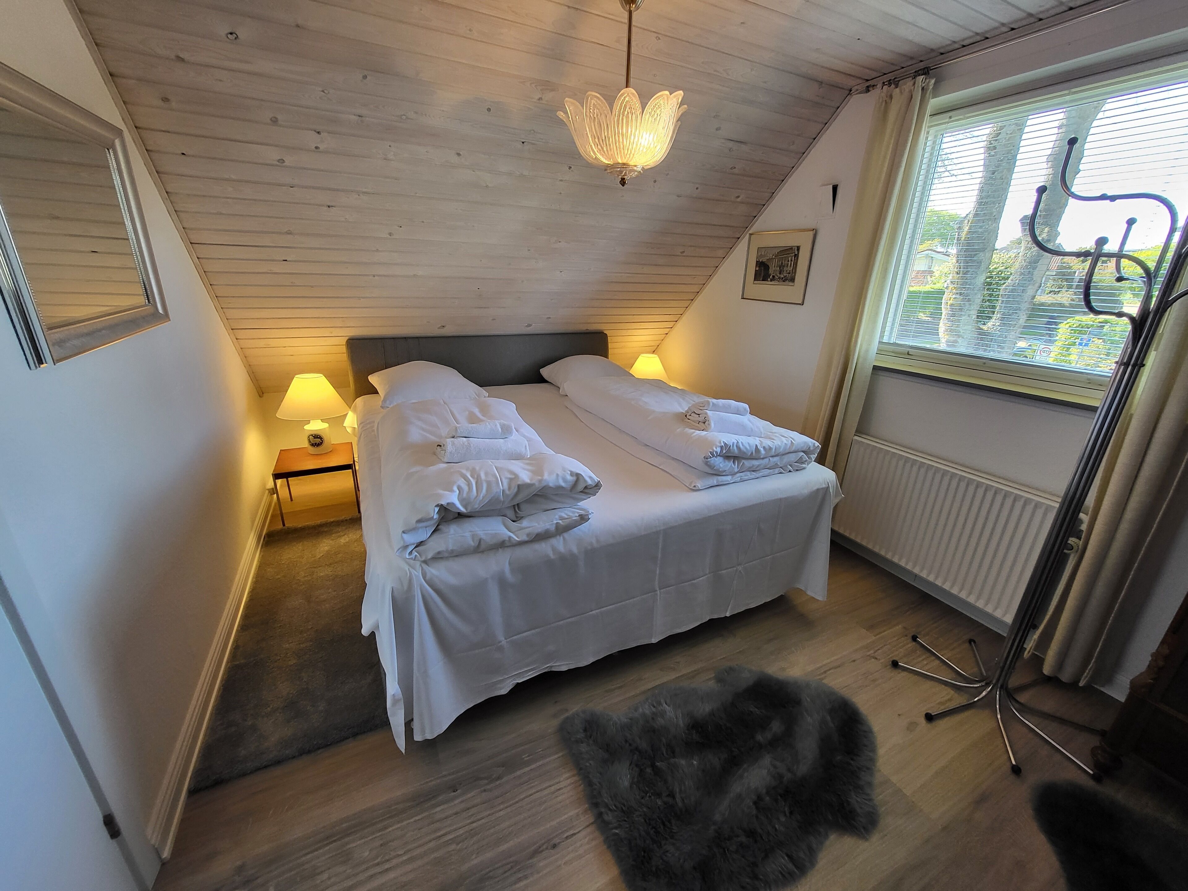 A bedroom with a wooden ceiling, a bed with white linens, a nightstand with a lamp, a radiator, and a window with blinds.