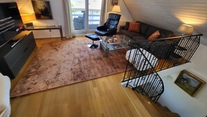 A cozy living room with a black leather chair, a glass coffee table, a brown rug, a sofa with orange cushions, and a staircase with a black railing. - Kollund Cottage (Krusaa)
