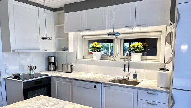 A modern kitchen with white cabinetry, a marble countertop, a Bosch dishwasher, and a sink with a window above it. - Kollund Cottage (Krusaa)
