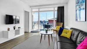 Ein modernes Wohnzimmer mit Flachbildfernseher, einer Couch mit bunten Kissen, einem Essbereich mit Tisch und Stühlen und großen Fenstern mit Blick auf die Stadt.