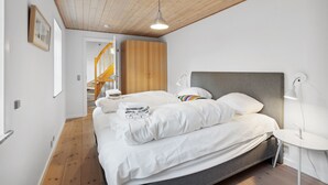 Un dormitorio moderno con techo de madera, cama con ropa de cama blanca, mesita de noche con lámpara y una escalera al fondo.