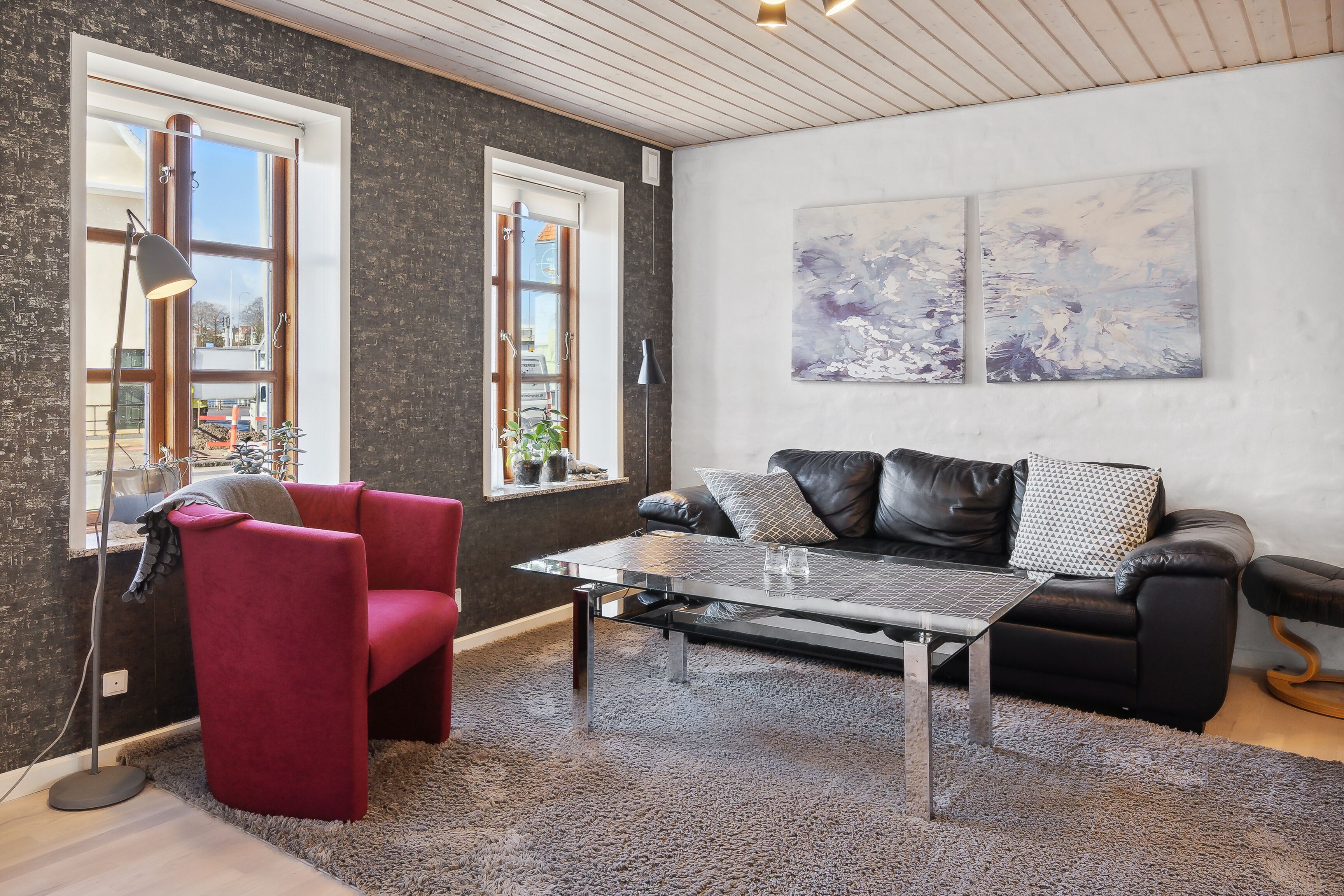 Un salon moderne avec un canapé en cuir noir, un fauteuil rouge et une table basse en verre. Il y a deux grandes fenêtres avec des rideaux, un lampadaire noir et des œuvres d’art abstraites accrochées au mur.