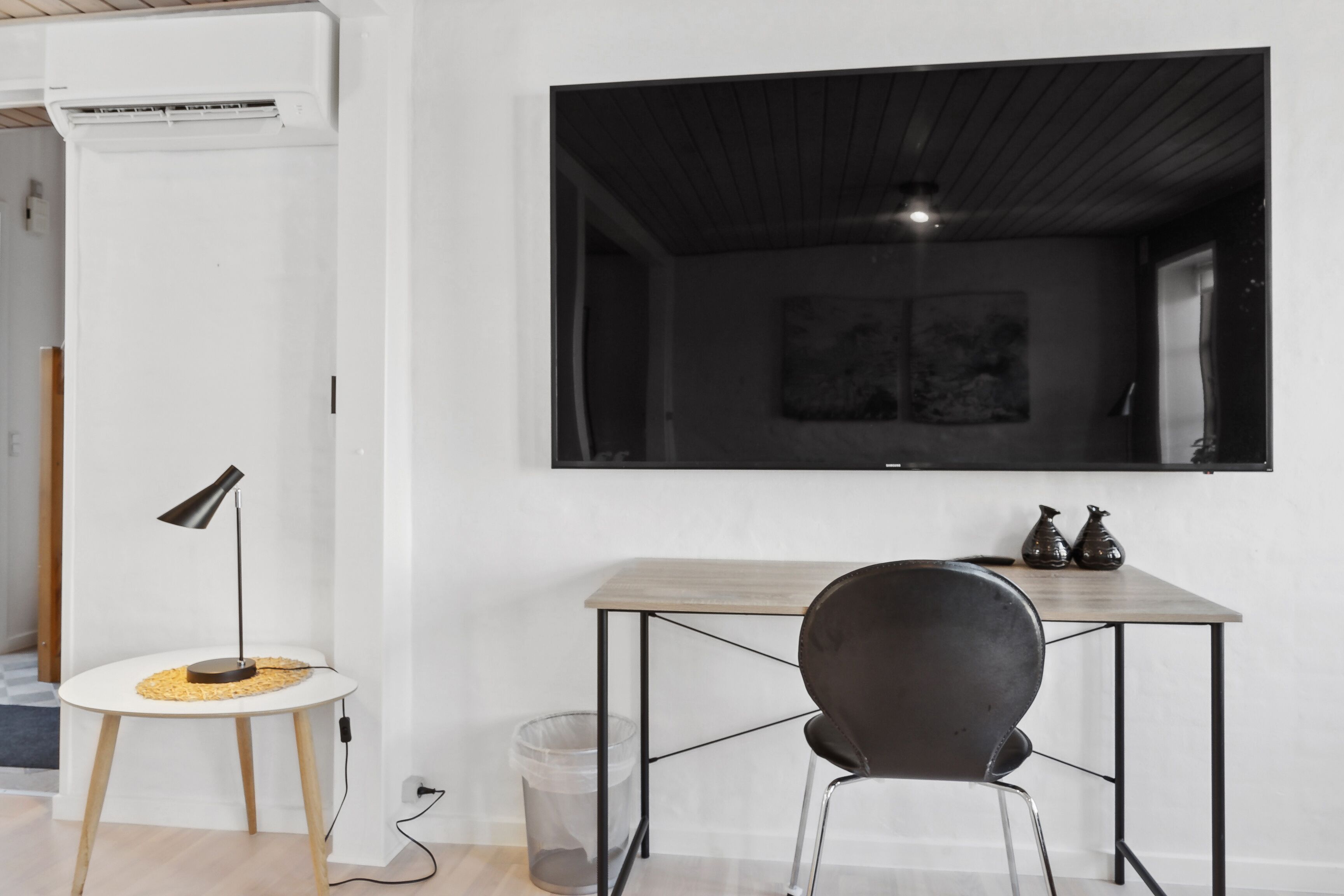 Een moderne kamer met houten vloer, een witte muur, een wandgemonteerde televisie, een klein rond tafeltje met een lamp, een zwarte stoel en een prullenbak.