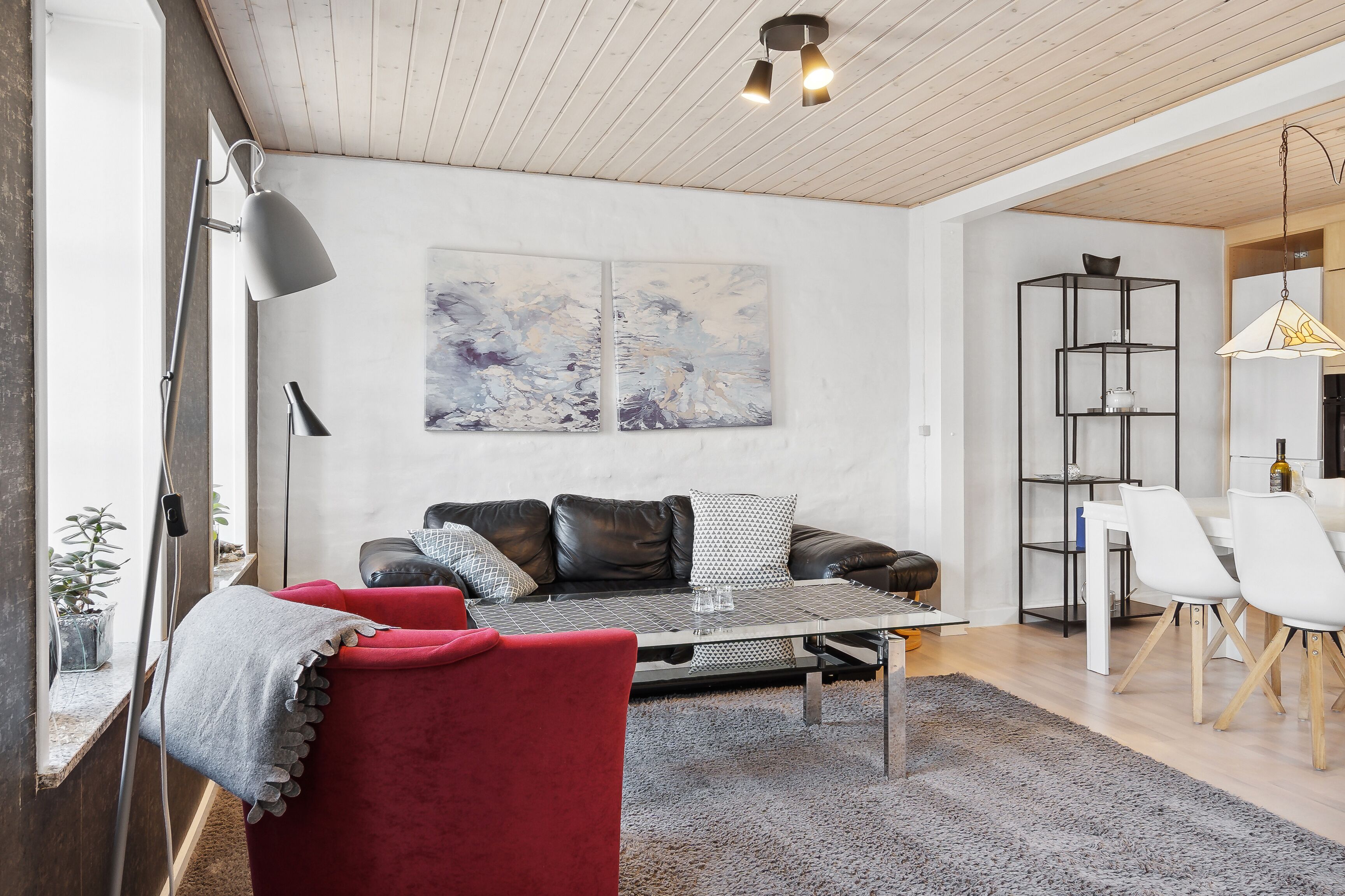 Una sala moderna con un sofá, una mesa de centro y un sillón rojo. Hay un cuadro en la pared y un comedor con una mesa y sillas.