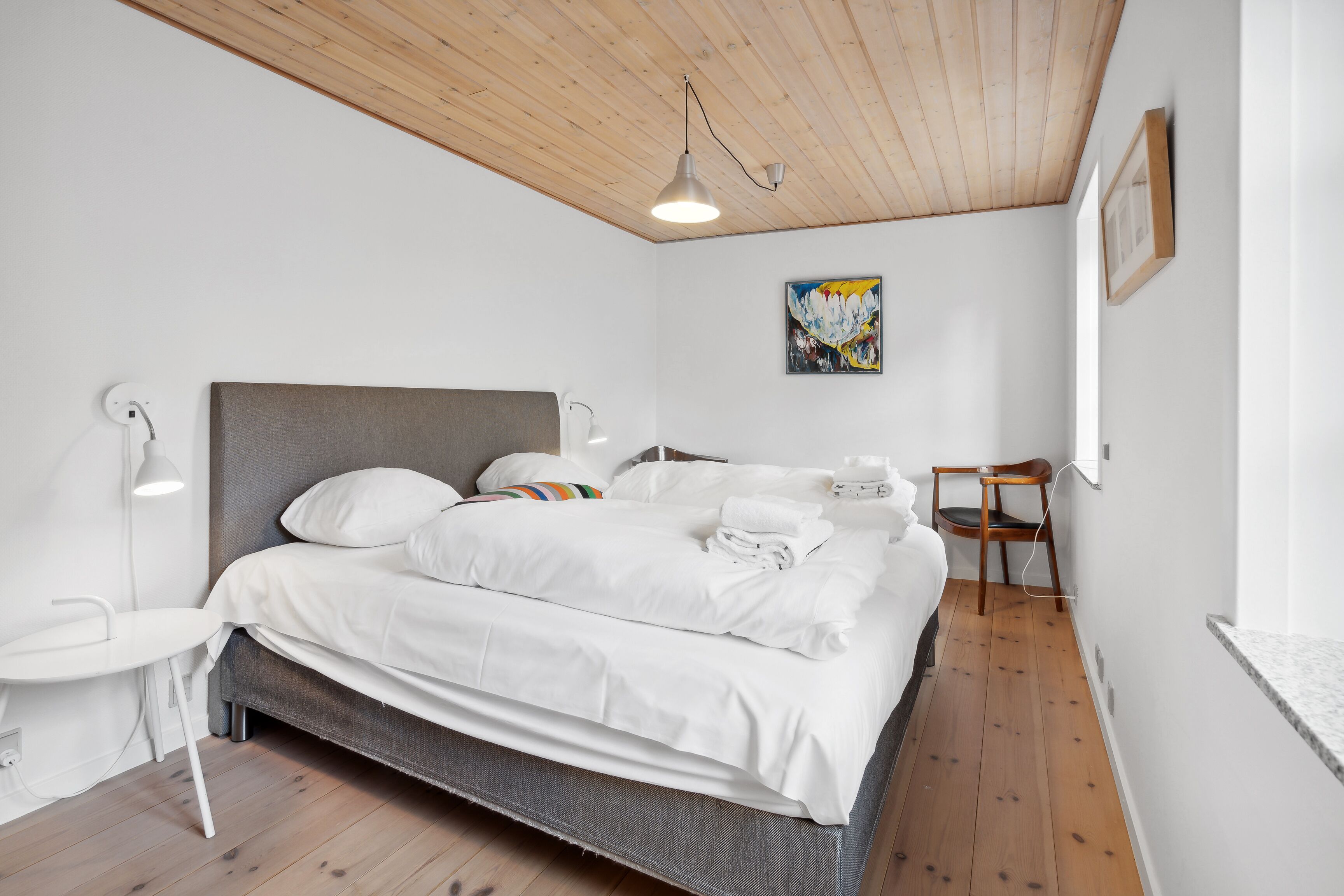 Uma cama bem arrumada com lençóis brancos, uma mesa de cabeceira com abajur e uma cadeira em um quarto com piso de madeira e teto inclinado.