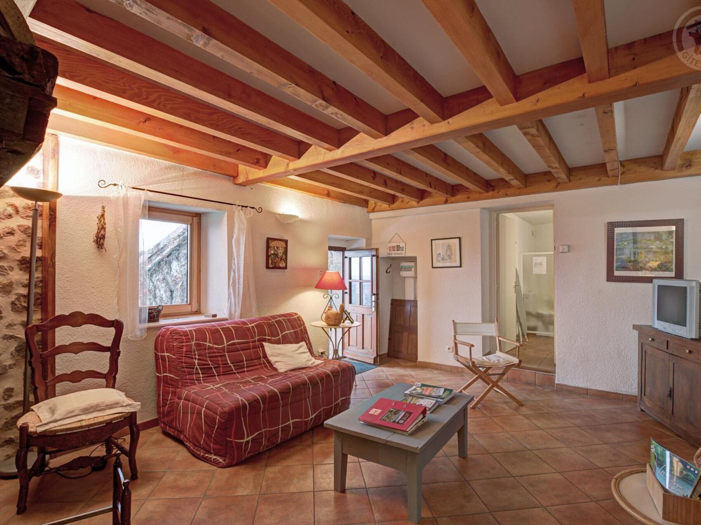 Une pièce chaleureuse avec un plafond en bois, un canapé rouge, une chaise en bois, une petite table avec des livres et un téléviseur posé sur un meuble.